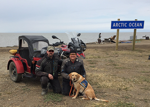 Michael and Judy sit with service dog Rasta in front of motorcycle with sidecar. The sign in background says "Arctic Circle". The gray ocean is in the background.